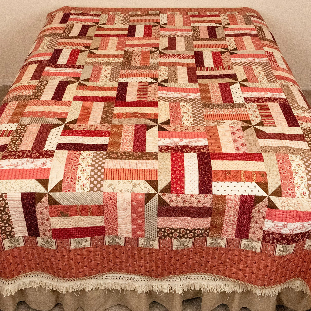 A patchwork quilted bedspread with a ruffled skirt, featuring a geometric pattern of red, orange, brown, and cream blocks, displayed on a bed in a bedroom corner.