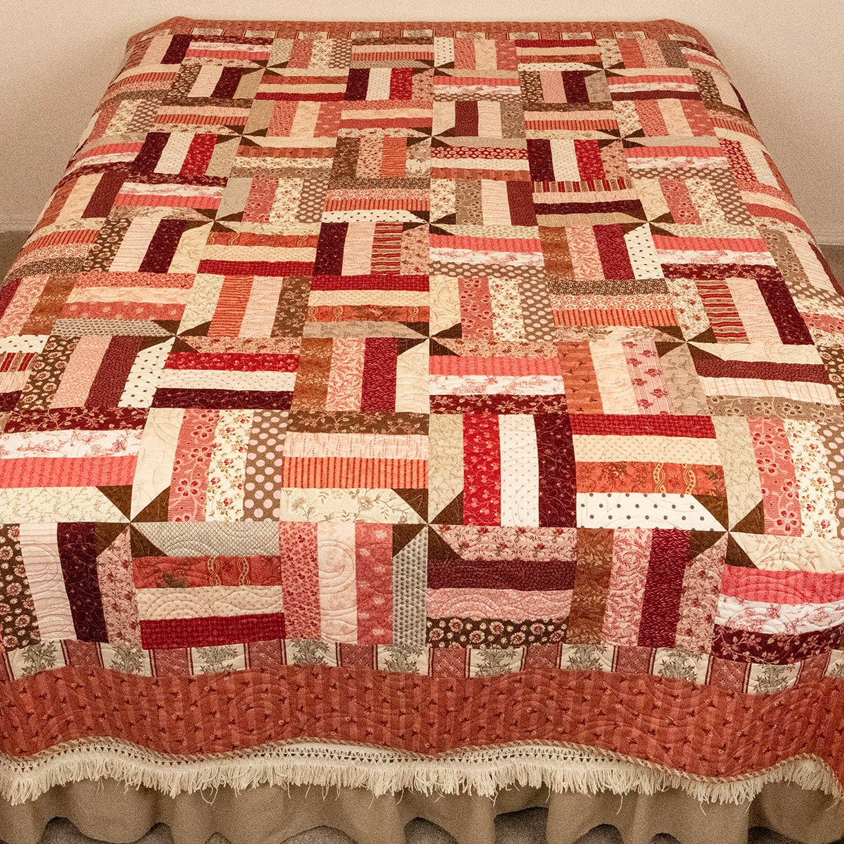 A patchwork quilted bedspread with a ruffled skirt, featuring a geometric pattern of red, orange, brown, and cream blocks, displayed on a bed in a bedroom corner.
