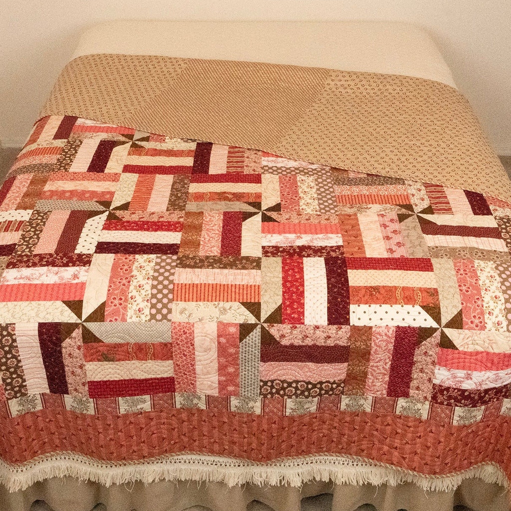 A patchwork quilted bedspread with a geometric block pattern in red, pink, brown, and beige, draped over a bed with a cream-colored ruffled skirt.