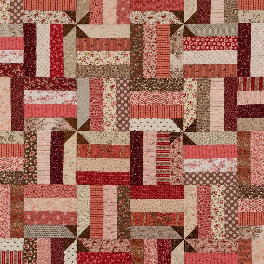 A patchwork quilt featuring a variety of red, pink, brown, and beige fabrics arranged in a geometric block pattern.