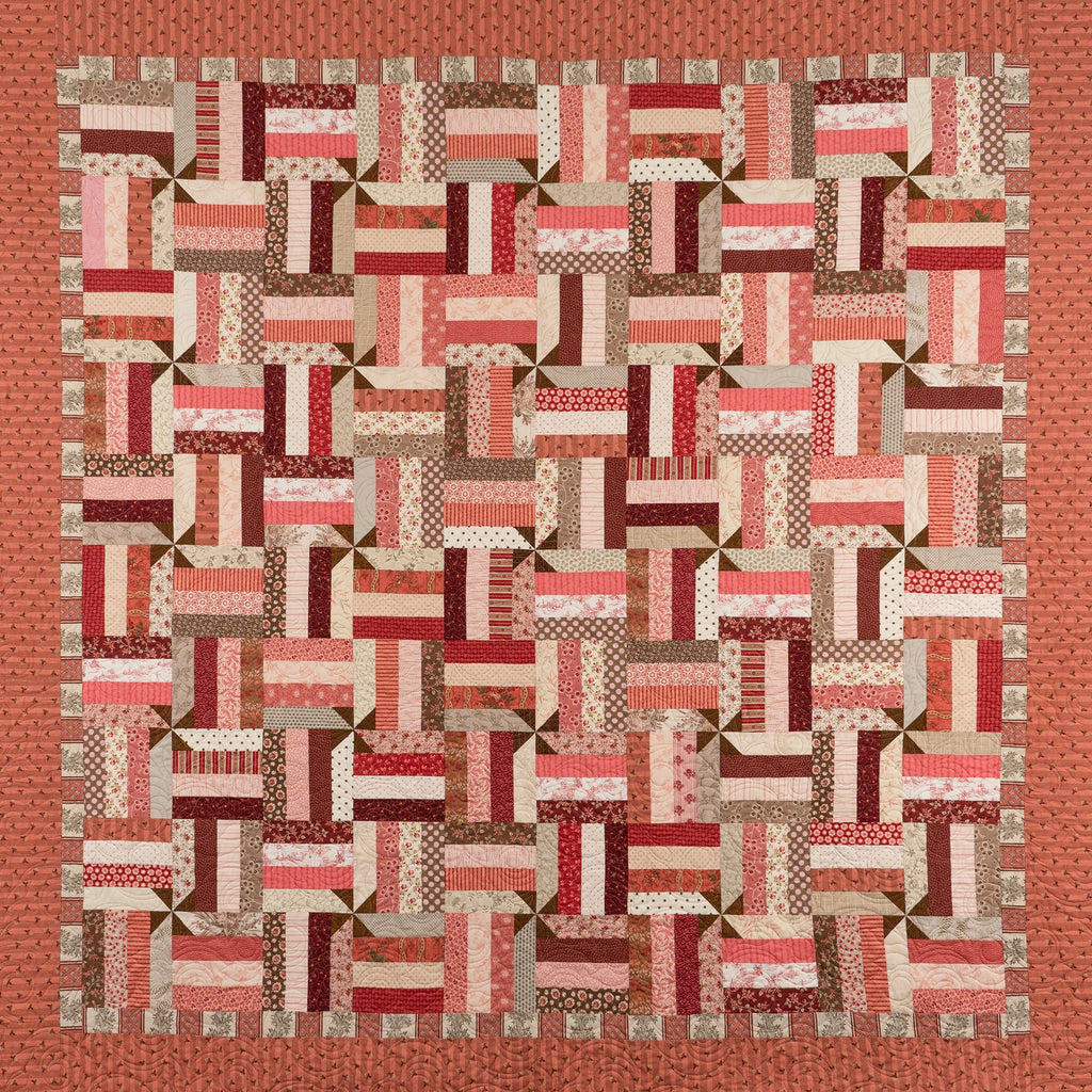 A square patchwork quilt with a geometric block pattern in shades of red, pink, brown, and cream, finished with a solid rust-orange border.