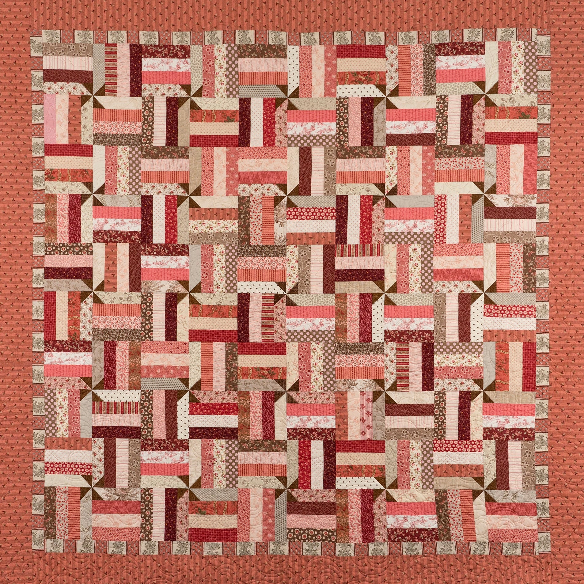 A square patchwork quilt with a geometric block pattern in shades of red, pink, brown, and cream, finished with a solid rust-orange border.