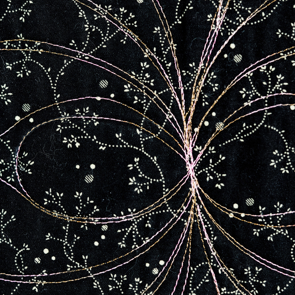 Black fabric with intricate embroidery featuring fine gold, pink, and white threads forming floral and leaf patterns radiating from a central point.