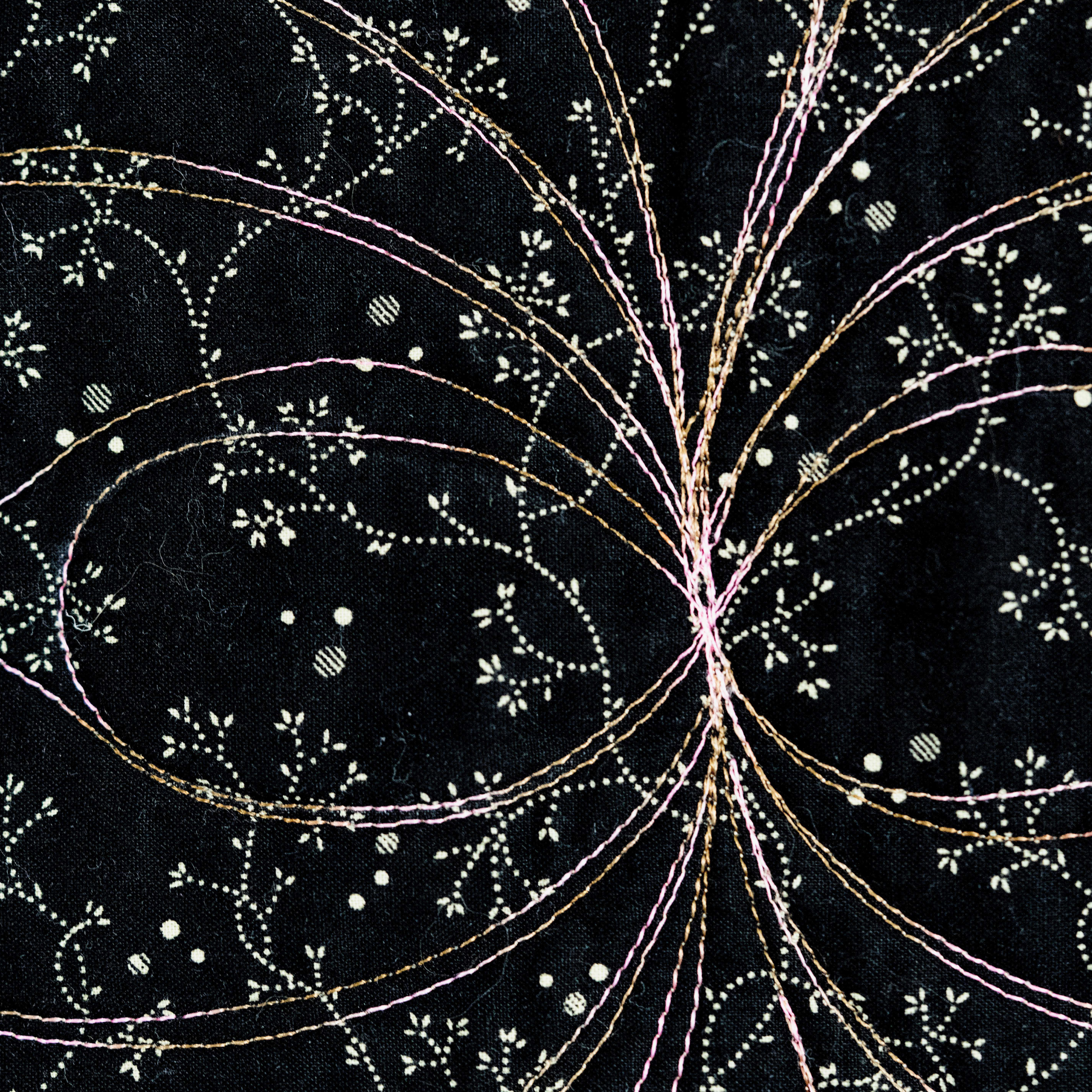Black fabric with intricate embroidery featuring fine gold, pink, and white threads forming floral and leaf patterns radiating from a central point.