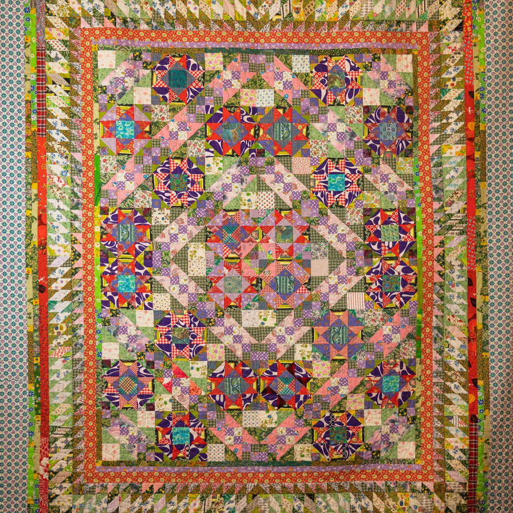 A large, intricate patchwork quilt with a complex geometric and floral pattern, featuring multiple colors including pink, purple, green, orange, and blue, bordered by a patterned fabric.
