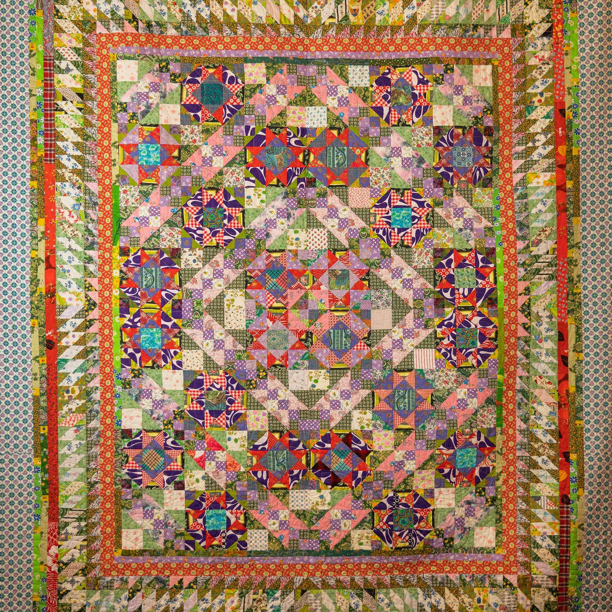 A large, intricate patchwork quilt with a complex geometric and floral pattern, featuring multiple colors including pink, purple, green, orange, and blue, bordered by a patterned fabric.