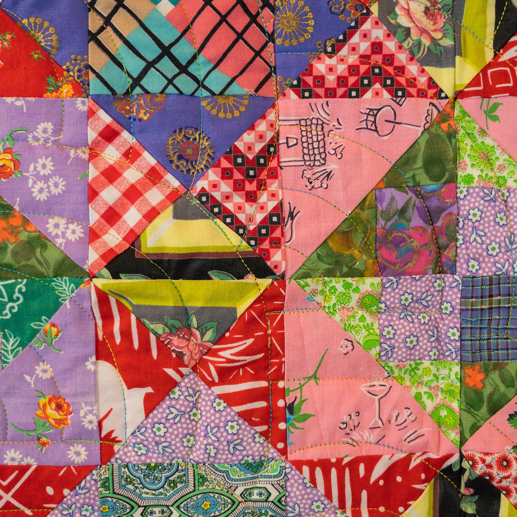 A colorful patchwork quilt featuring various fabric patterns including gingham, florals, plaid, and stripes in pink, red, green, purple, blue, and yellow.
