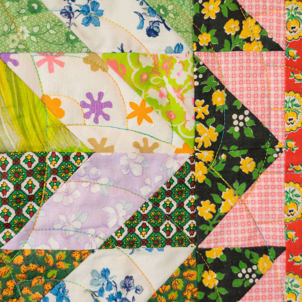 A close-up of a colorful quilted fabric panel with multiple patterned sections including floral, geometric, and checkered designs in green, pink, yellow, orange, purple, and red.