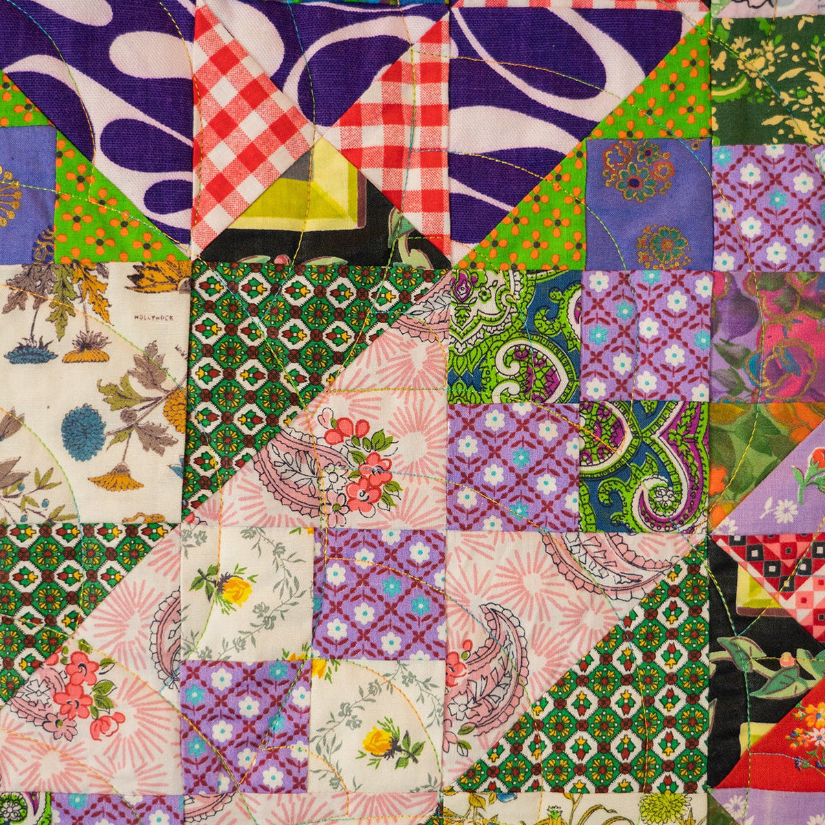 A square patchwork quilt featuring a variety of colorful fabric patterns including gingham, paisley, floral prints, and abstract designs in purple, green, pink, red, and white.