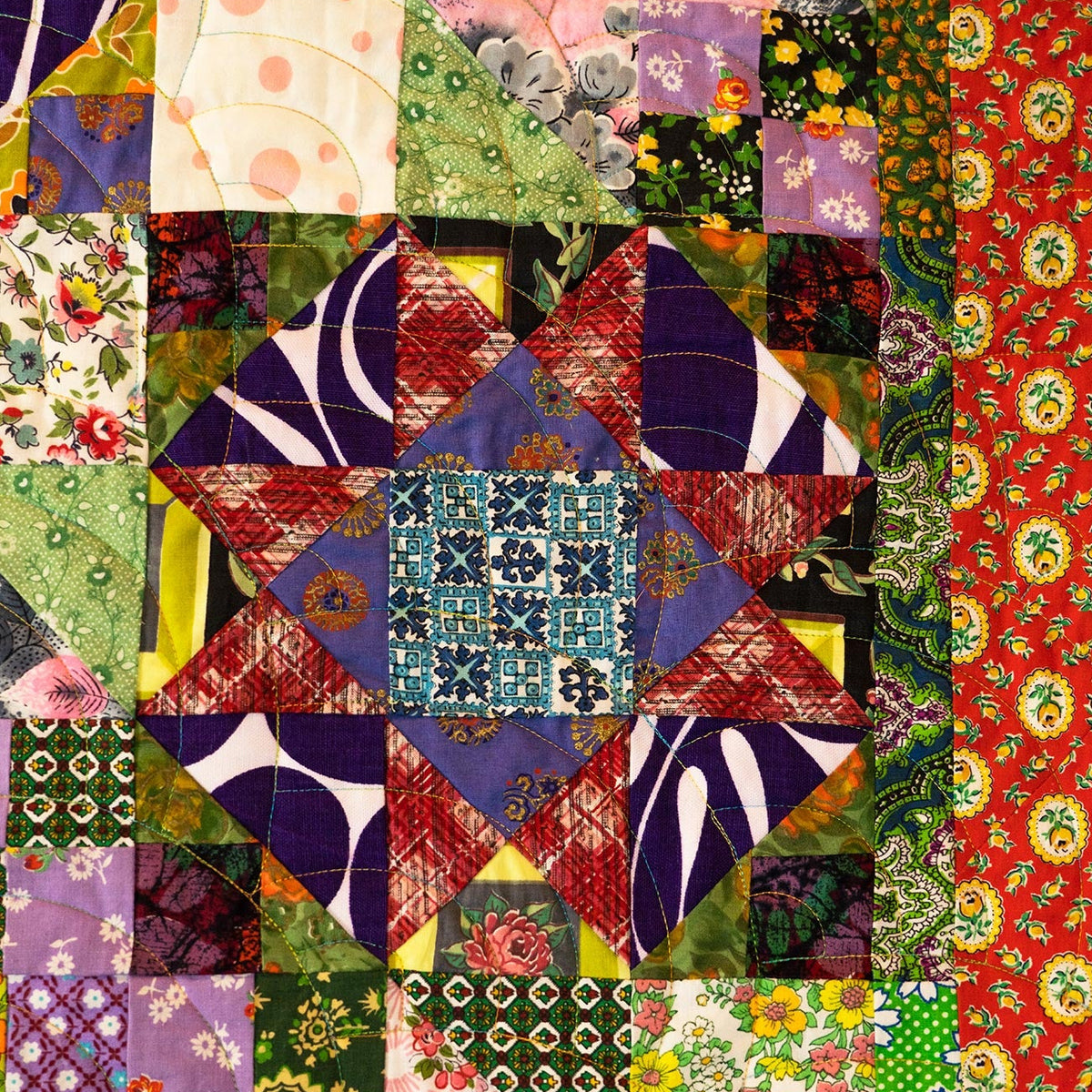 A colorful patchwork quilt featuring a central starburst design with geometric and floral patterns, surrounded by striped and floral fabric sections in red, green, purple, pink, and blue.