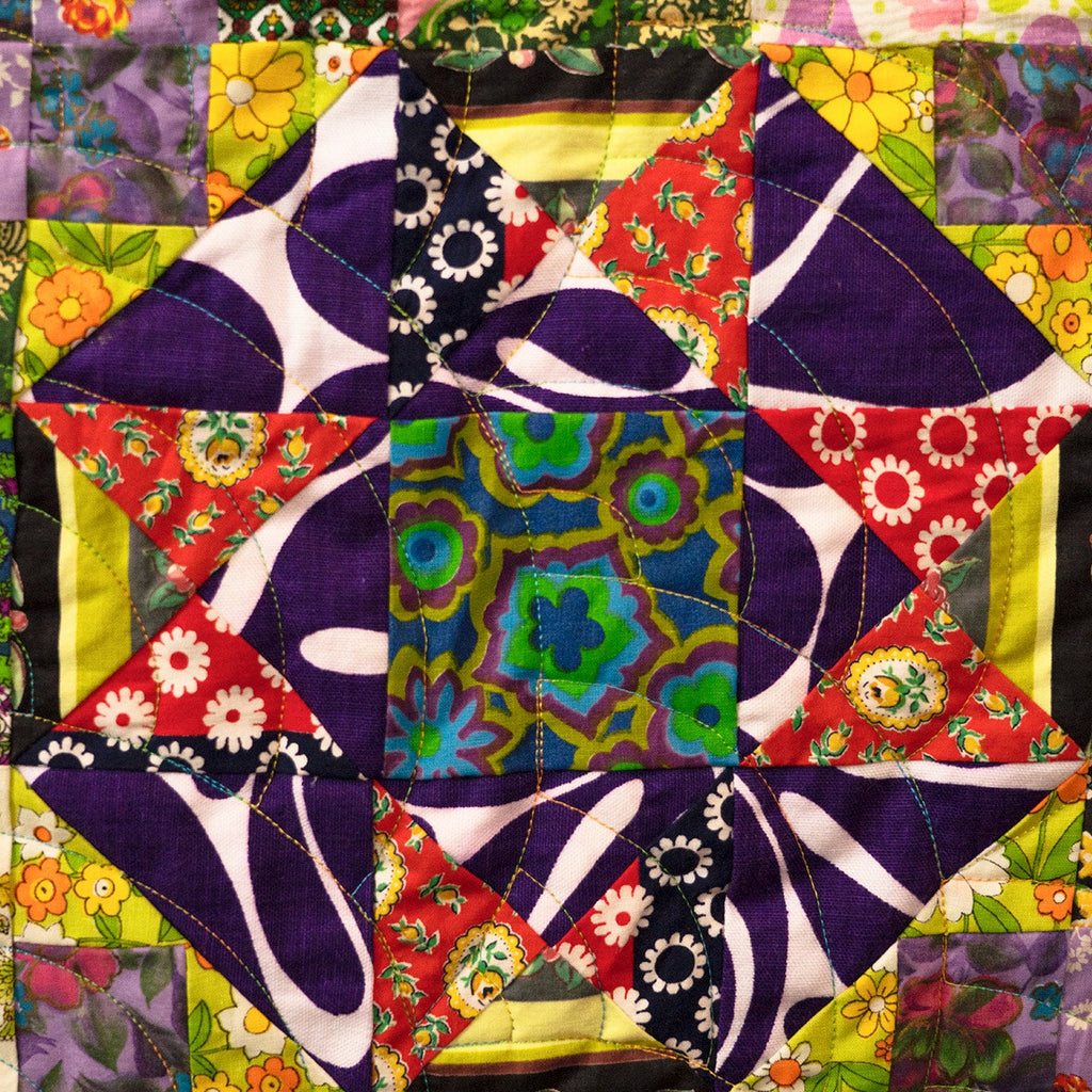 A colorful patchwork quilt featuring a central star-like geometric design with purple, green, blue, red, and yellow fabrics, surrounded by a border of floral and patterned quilt blocks.