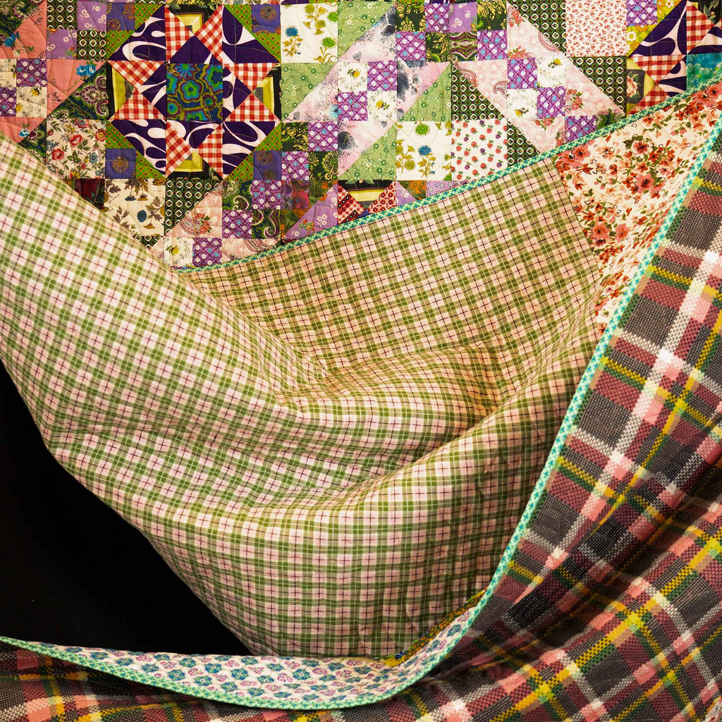 A multicolor patchwork quilt with a geometric starburst pattern on the top half and a plaid reverse side, draped to show both sides against a black background.