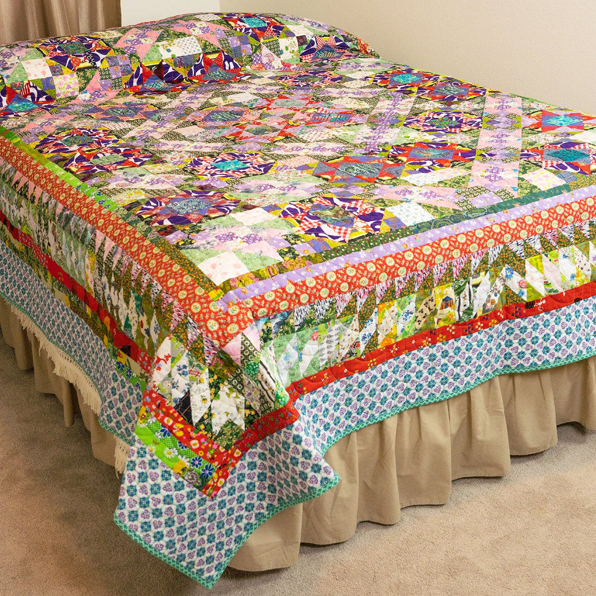 A colorful patchwork quilt with a floral and geometric pattern, styled on a bed with a beige ruffled bed skirt in a bedroom setting.