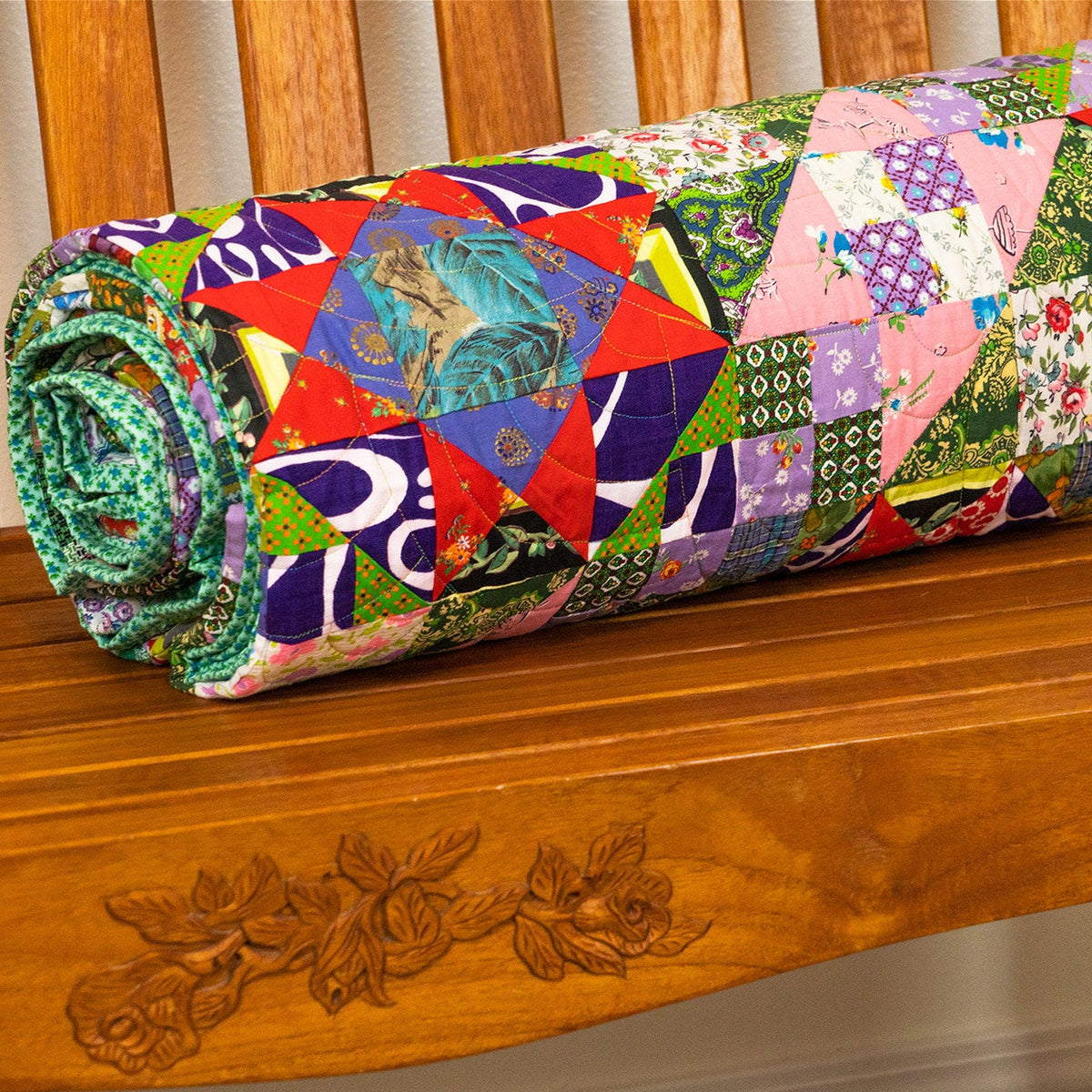 A colorful patchwork quilt rolled up and resting on a wooden bench.