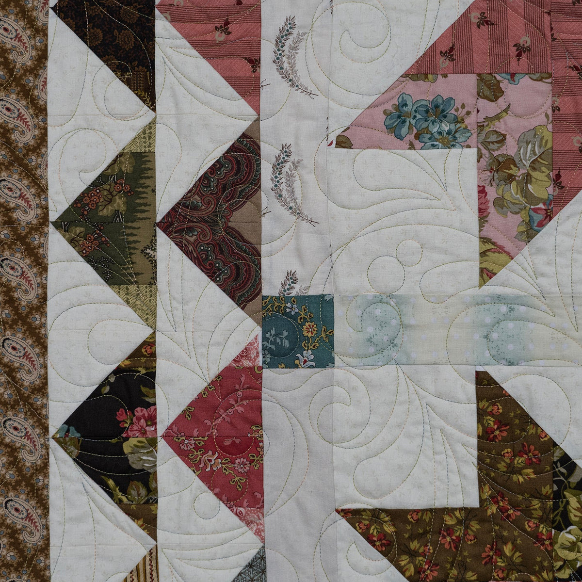 A close-up of a patchwork quilt featuring various fabric patterns including paisley, floral, and solid colors, with white quilting stitching.