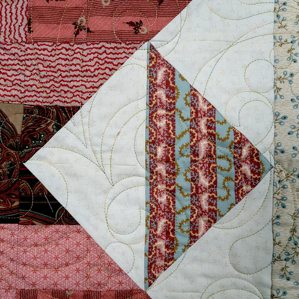 A close-up of a patchwork quilt featuring multiple fabric panels with different patterns, including paisley, wavy lines, florals, and geometric designs in pink, red, brown, beige, and light blue. The quilt has visible quilting stitches and is photographed filling the frame.