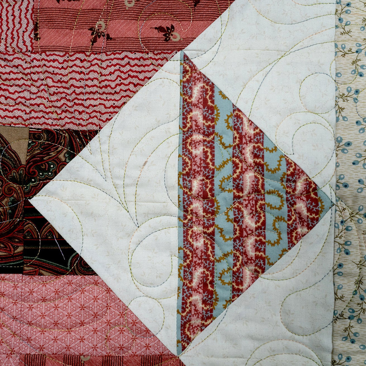 A close-up of a patchwork quilt featuring multiple fabric panels with different patterns, including paisley, wavy lines, florals, and geometric designs in pink, red, brown, beige, and light blue. The quilt has visible quilting stitches and is photographed filling the frame.
