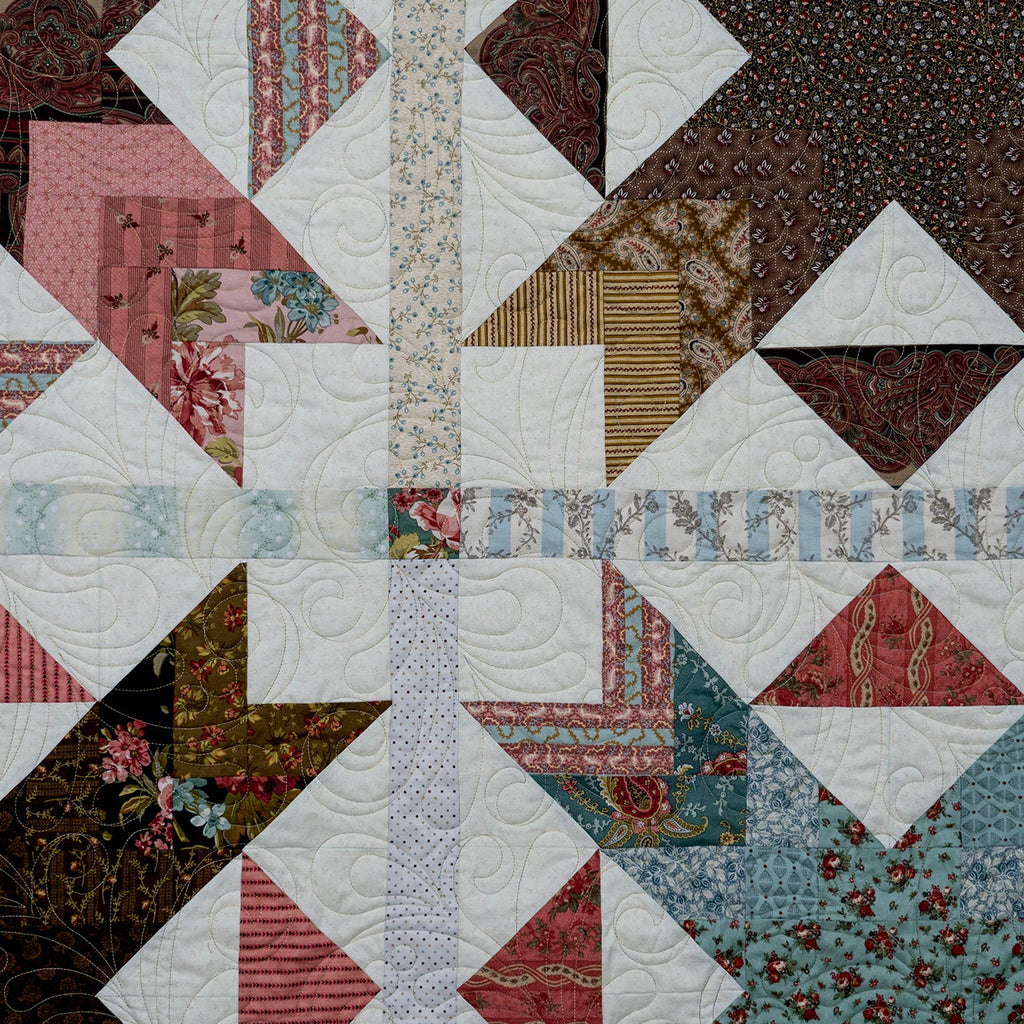 A close-up of a patchwork quilt featuring geometric star and triangle blocks in various fabrics, including floral prints, stripes, and solid colors like pink, brown, white, and light blue.