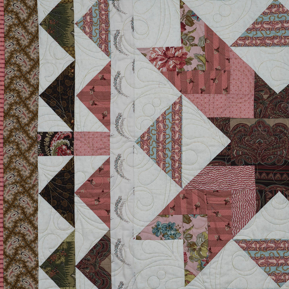 A square quilted textile featuring a geometric starburst pattern made from pink, dusty rose, brown, white, and small teal floral patterned fabric triangles, with a decorative border on the left side.