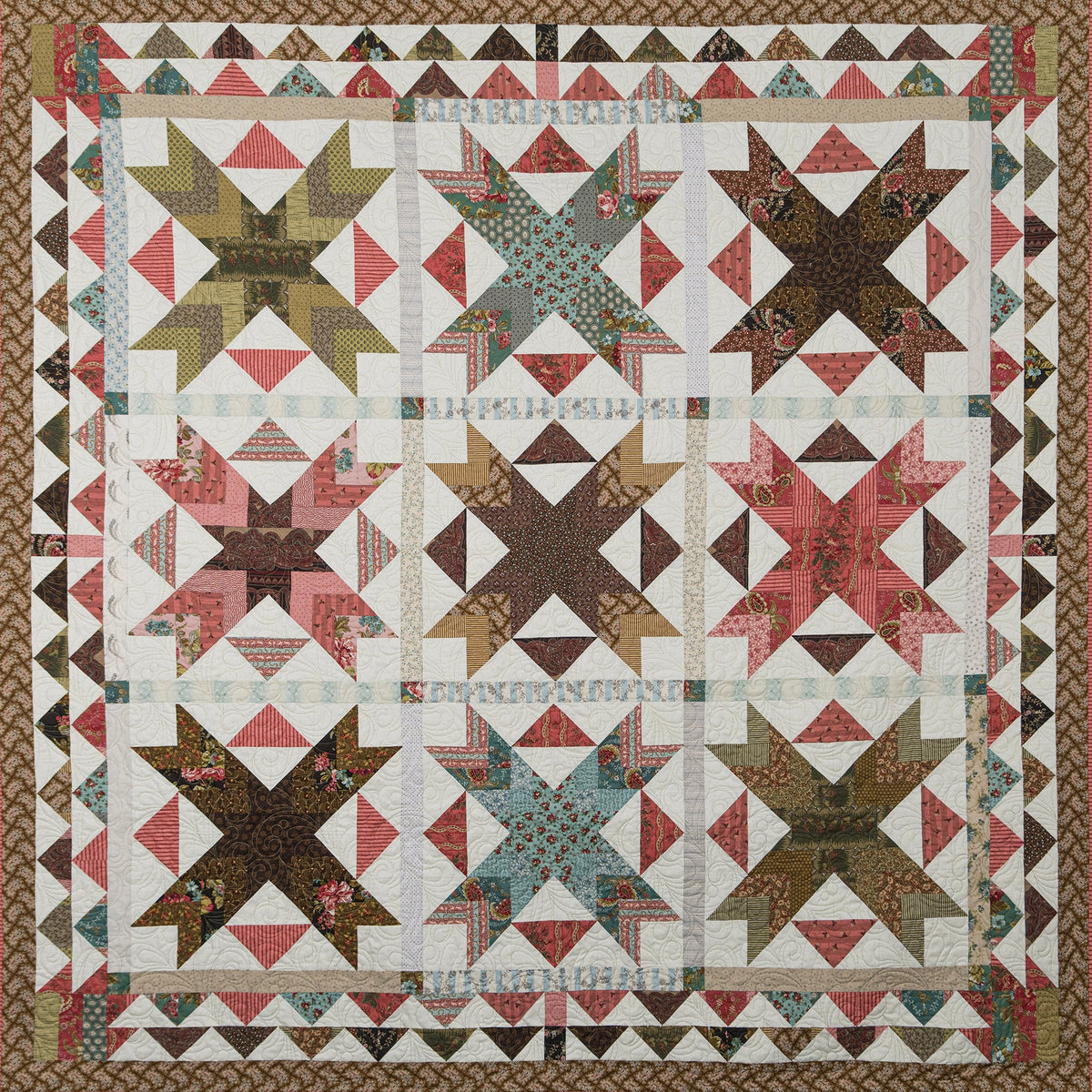 A vintage-style quilted textile featuring a geometric and triangle pattern in muted tones of pink, brown, olive green, and cream, finished with a decorative patterned border.