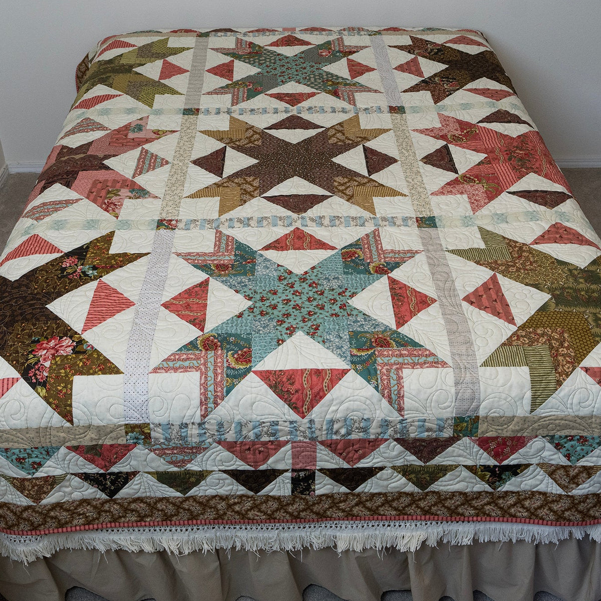 A large quilted patchwork bedspread with a ruffled skirt, featuring a geometric and triangle pattern in cream, brown, red, and teal, shown on a bed in a bedroom.