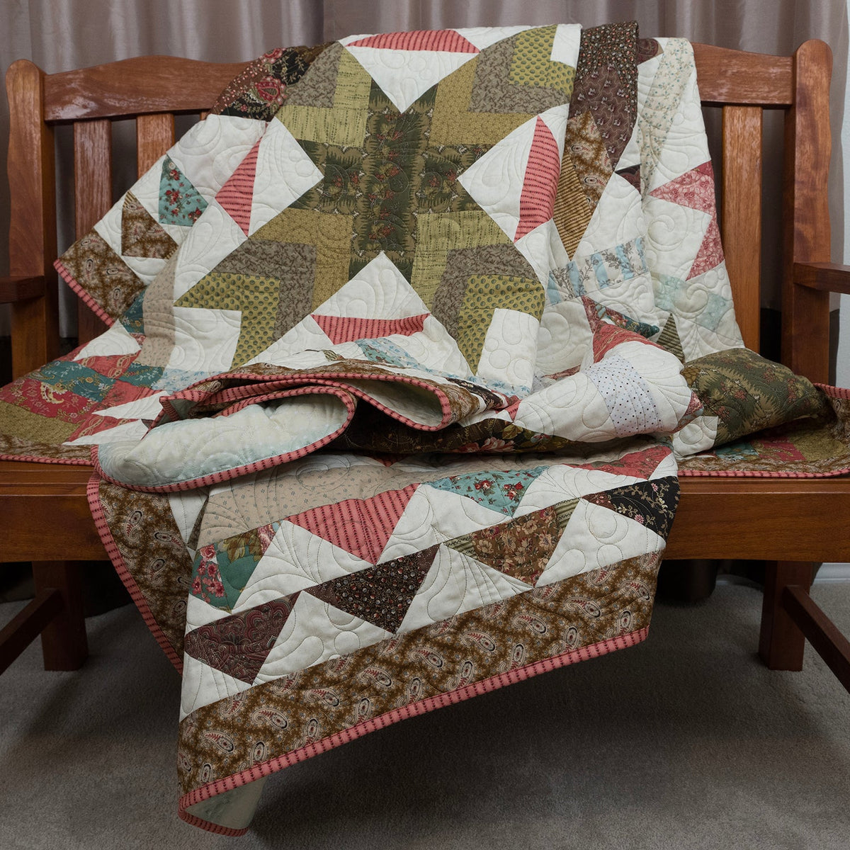 A patchwork quilt with geometric triangles in olive green, brown, red, and white, draped over a wooden bench in an indoor setting.