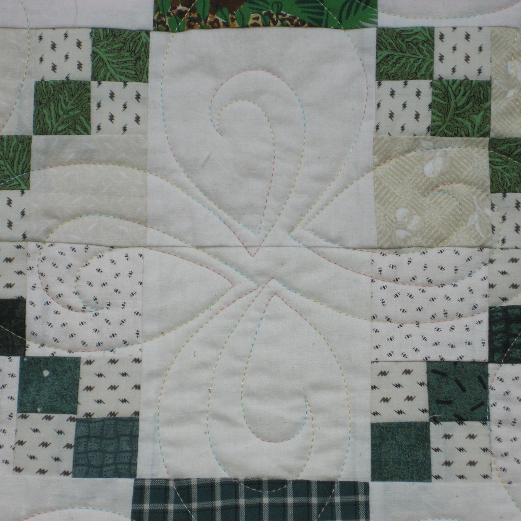 A close-up of a quilted patchwork fabric featuring a central white block with swirling stitched quilting, surrounded by green and white patterned squares.