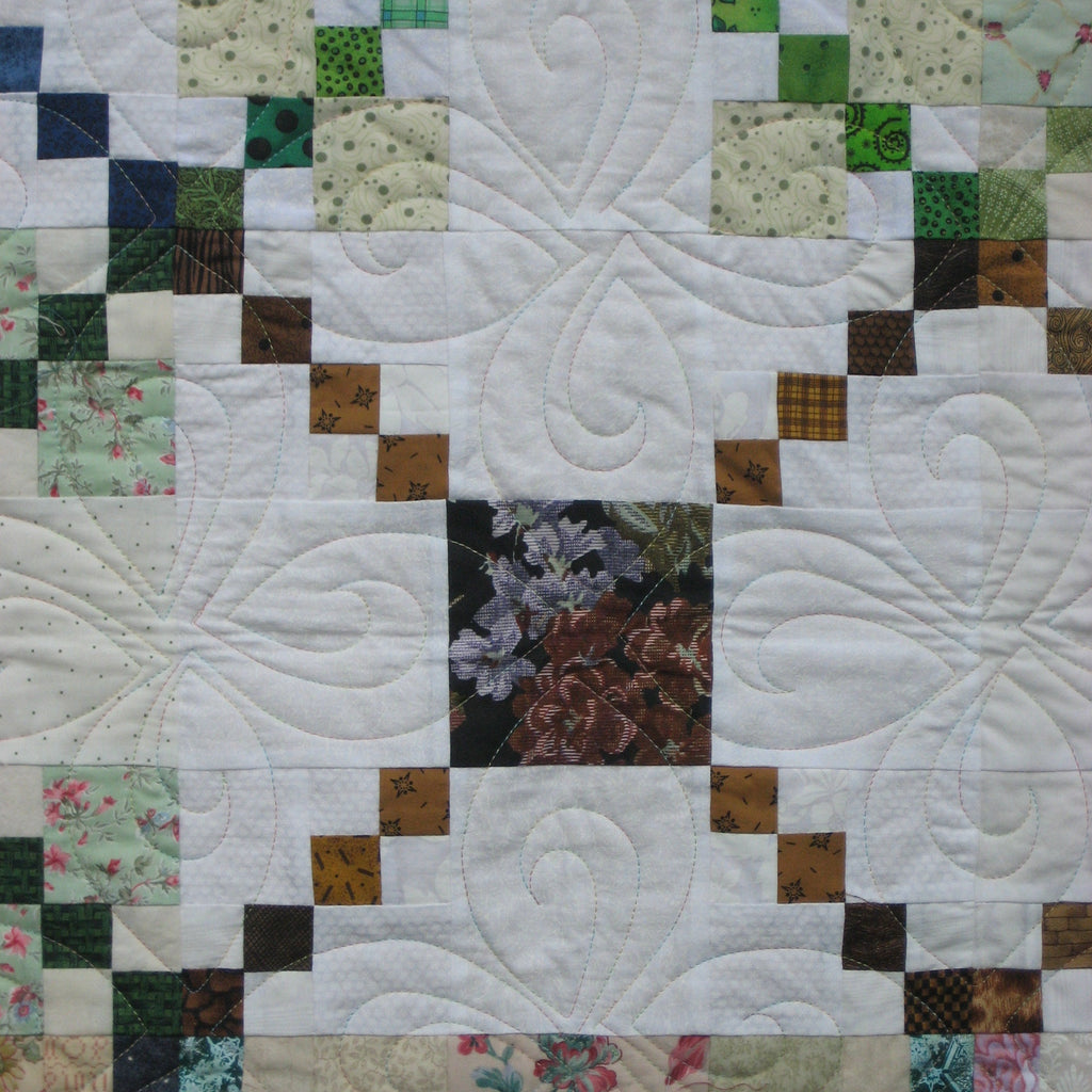A close-up of a quilted patchwork fabric featuring a white background with swirling stitched patterns, bordered by green, brown, floral, and plaid quilted squares.