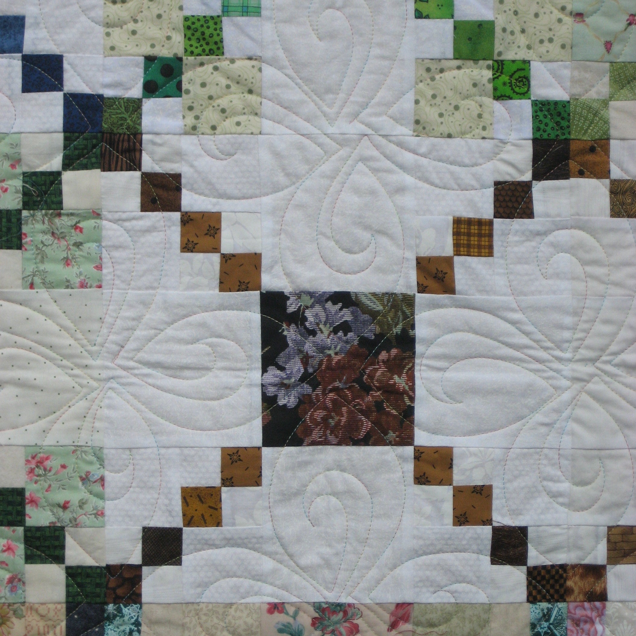 A close-up of a quilted patchwork fabric featuring a white background with swirling stitched patterns, bordered by green, brown, floral, and plaid quilted squares.