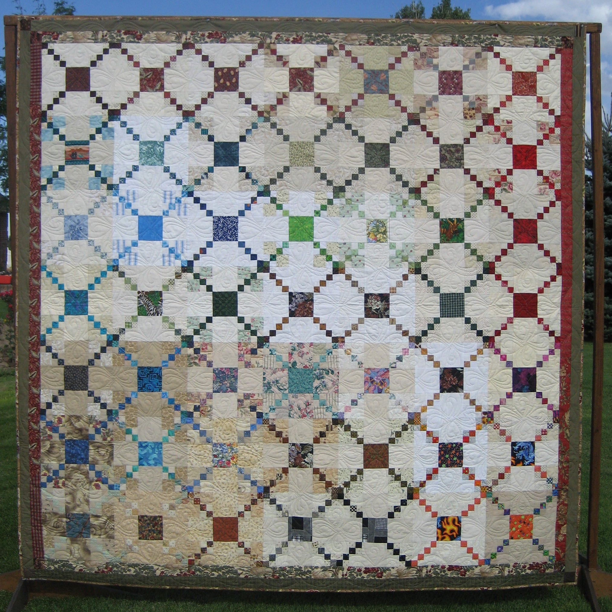 A large patchwork quilt displayed outdoors on a wooden frame against a blue sky with clouds and green grass. The quilt features a geometric diamond pattern with cream, beige, brown, red, blue, green, and black fabrics.