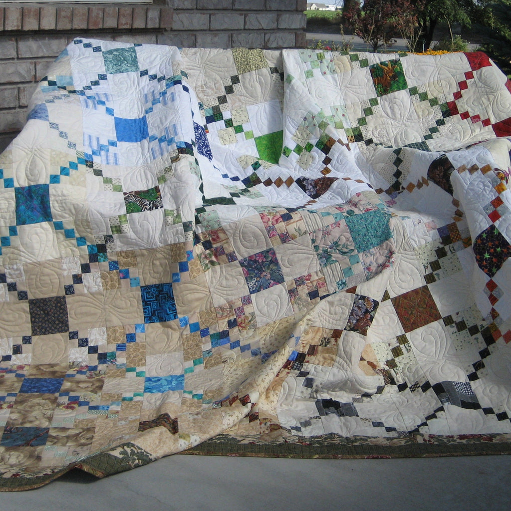 A large patchwork quilt draped over a bench on an outdoor patio, featuring a geometric diamond pattern in cream, blue, green, red, and brown with a decorative border.