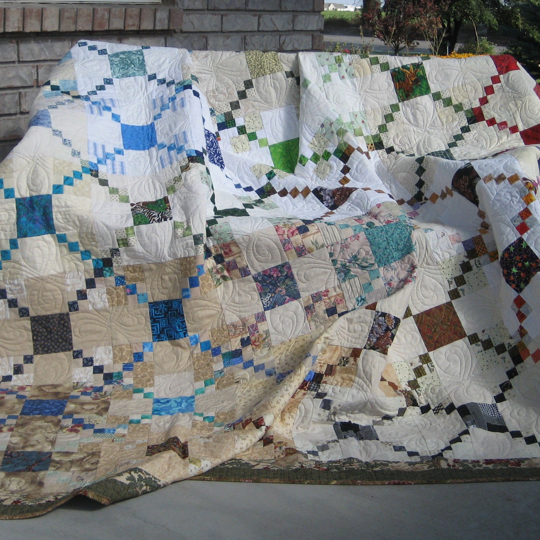 A large patchwork quilt draped over a bench on an outdoor patio, featuring a geometric diamond pattern in cream, blue, green, red, and brown with a decorative border.