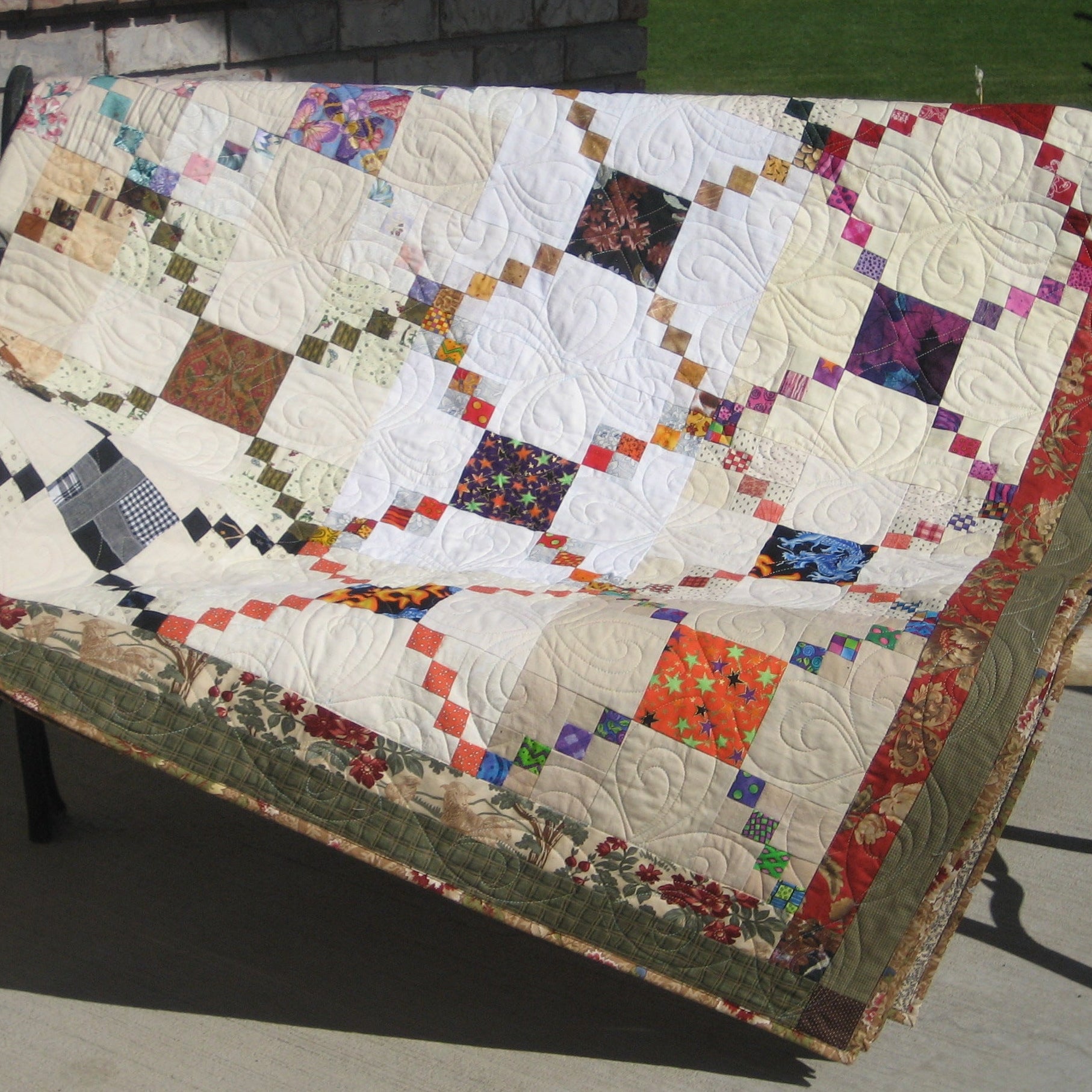 A handmade patchwork quilt draped over a black metal garden bench on a concrete patio, with a brick wall and green grass in the background.