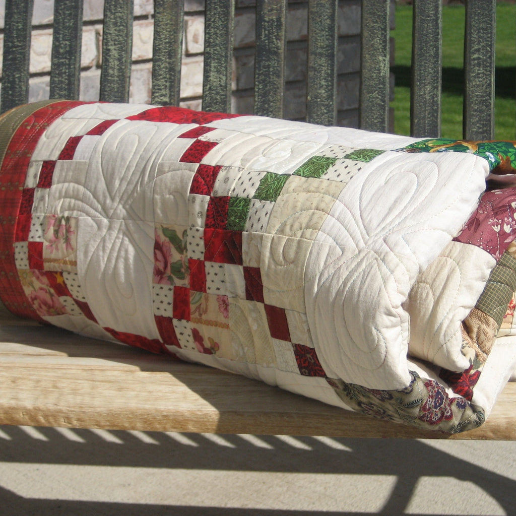 A summer quilt rolled-up with a red, green, and cream patchwork design, resting on a wooden bench in outdoor sunlight.