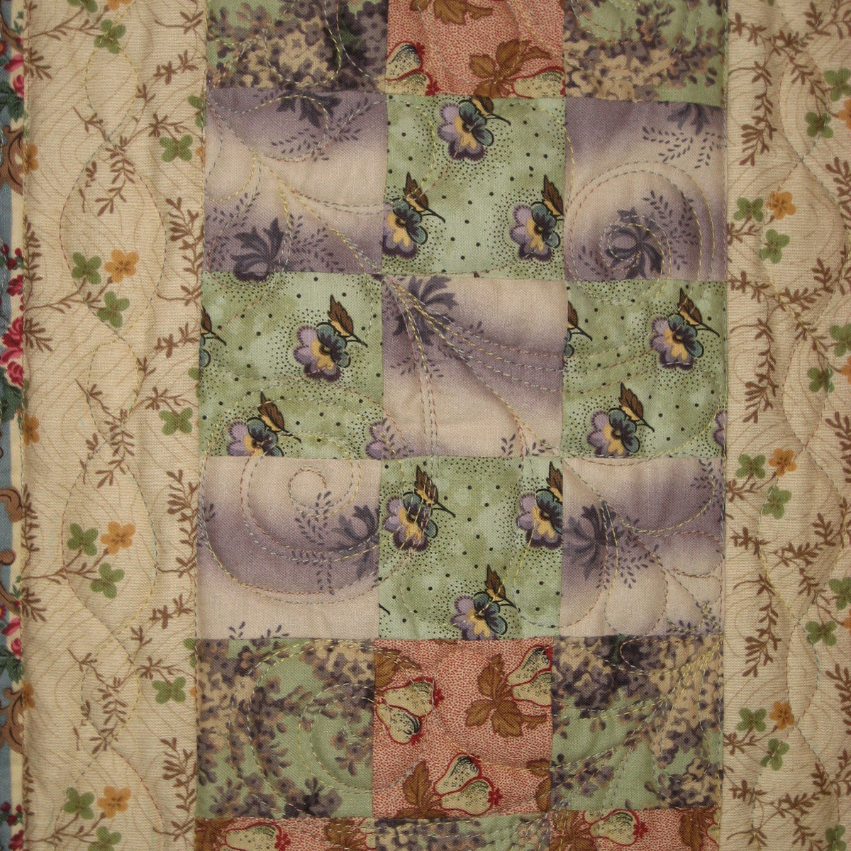 A close-up of a patchwork quilt featuring various floral patterned fabric squares in muted shades of green, pink, purple, and beige, bordered by a floral printed fabric.