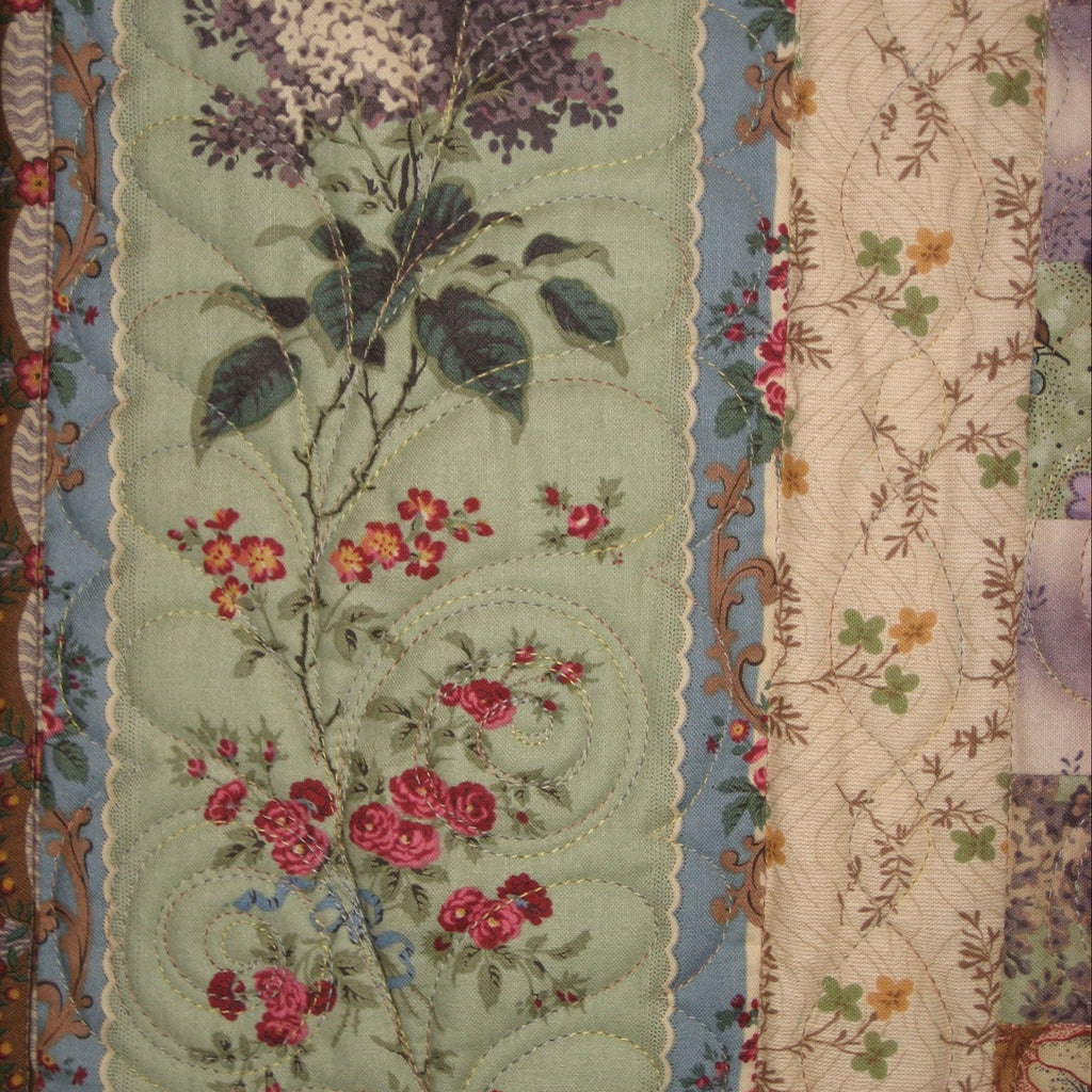 Close-up of a quilted textile featuring floral patterns, including a central pale green panel with purple flowers and pink roses, bordered by blue and cream sections with additional botanical motifs.