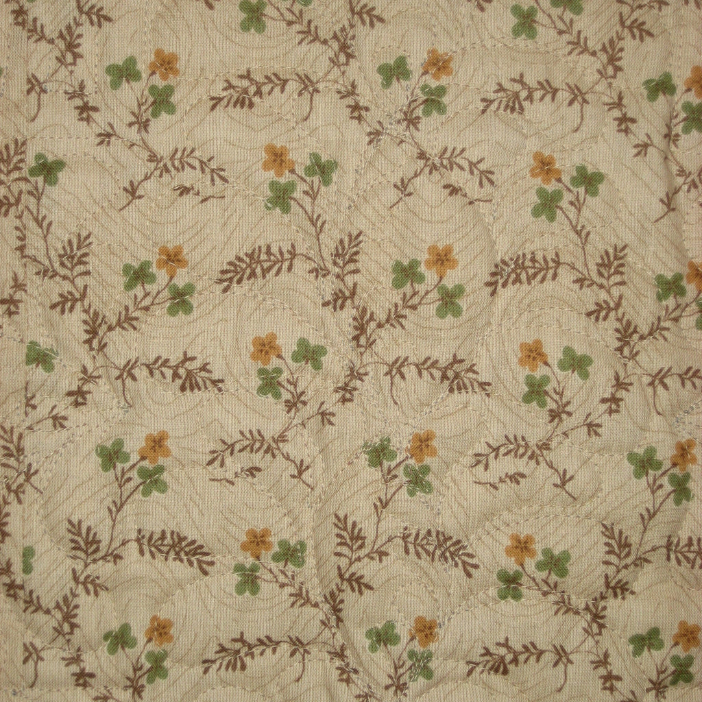 A close-up of a quilted fabric featuring a repeating pattern of small orange flowers, green clover-like leaves, and brown leafy vines over a cream background.