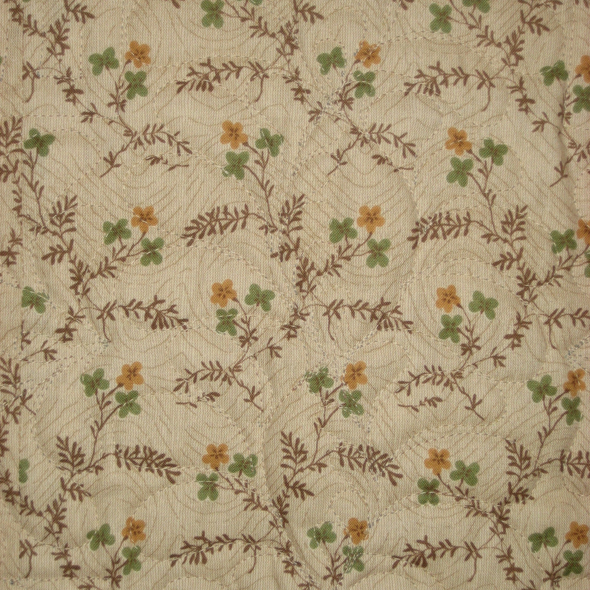 A close-up of a quilted fabric featuring a repeating pattern of small orange flowers, green clover-like leaves, and brown leafy vines over a cream background.