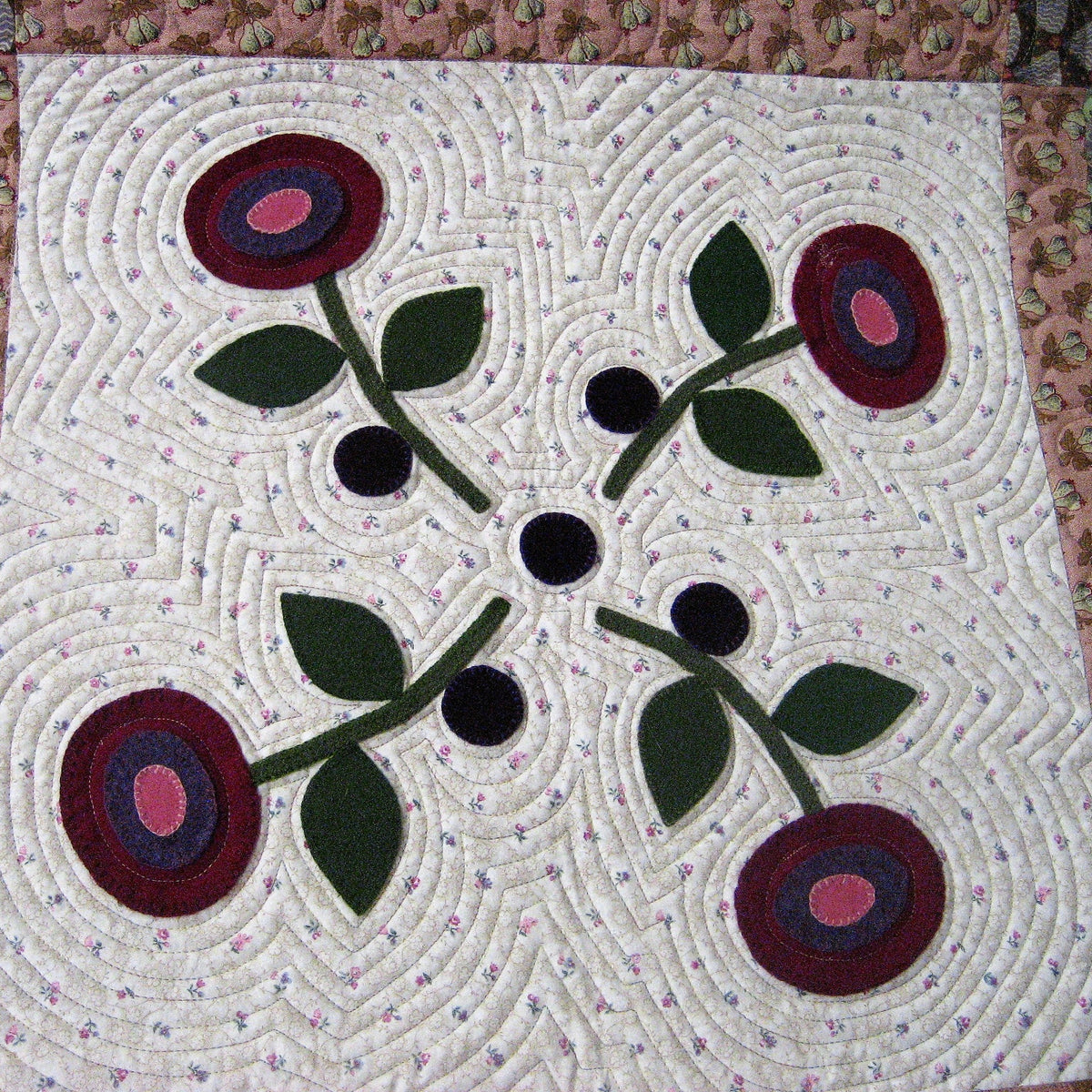 A square quilted textile panel featuring a symmetrical floral design with four burgundy and pink roses, green leaves, and black centers on a cream background, surrounded by a pink patterned border.